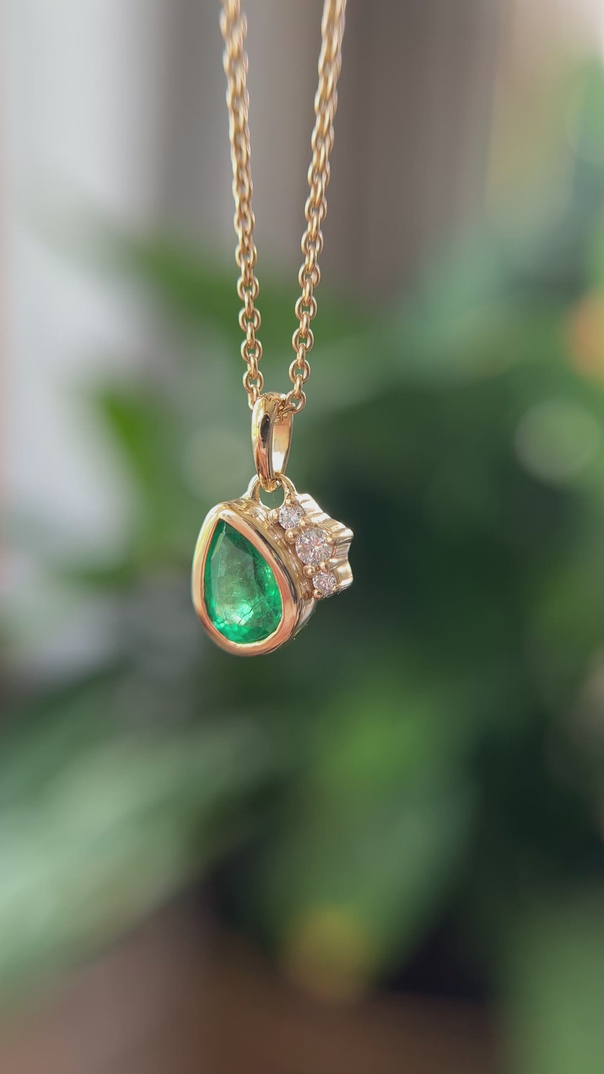 14k yellow gold pendant, set with a pear-shaped emerald and natural diamond accents. The pendant is hanging from a chain, in front of a green blurry background