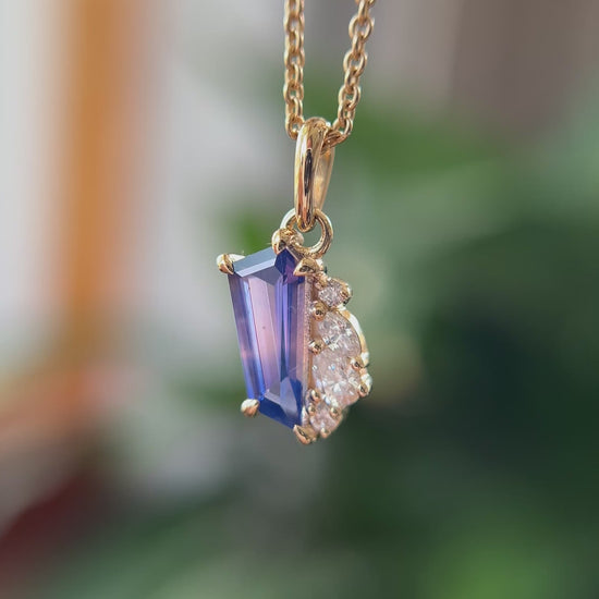 14k yellow gold pendant, set with a bi-color blue to pink sapphire and natural diamond accents. The pendant is hanging from a chain, in front of a green blurry background