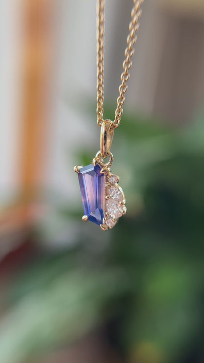 14k yellow gold pendant, set with a bi-color blue to pink sapphire and natural diamond accents. The pendant is hanging from a chain, in front of a green blurry background