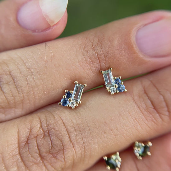 Cluster stud pair of earrings set with blue gemstones, held between fingers besides cluster stud pair of earrings set with green gemstones. 