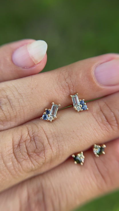 Cluster stud pair of earrings set with blue gemstones, held between fingers besides cluster stud pair of earrings set with green gemstones. 