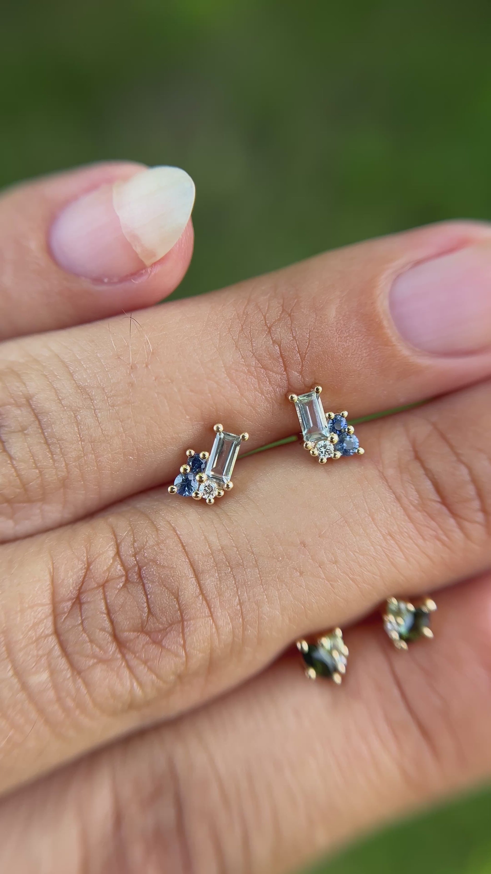 Cluster stud pair of earrings set with blue gemstones, held between fingers besides cluster stud pair of earrings set with green gemstones. 