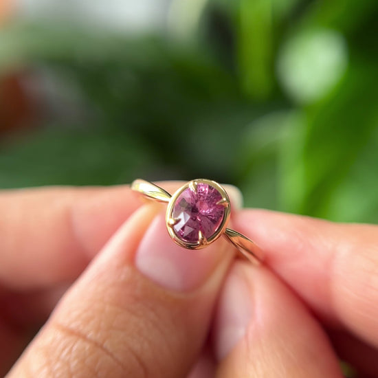 1.20ct oval orangy-pink sapphire solitaire engagement ring in 14k yellow gold with gold frame eagle-claw setting, held in hands showing gemstone colour and faceting