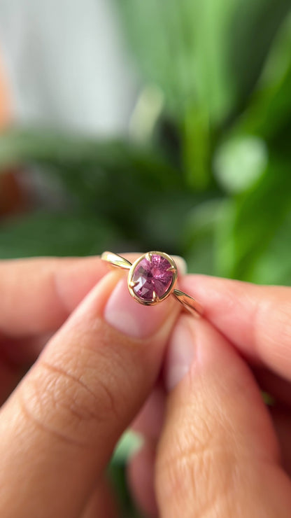 1.20ct oval orangy-pink sapphire solitaire engagement ring in 14k yellow gold with gold frame eagle-claw setting, held in hands showing gemstone colour and faceting
