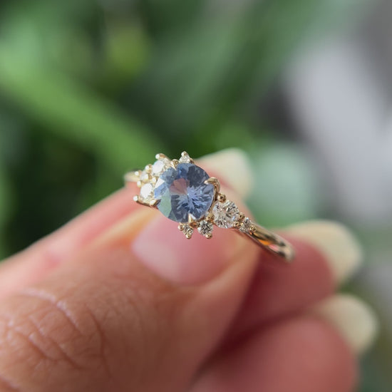 0.82ct round unheated cornflower blue Sri Lankan sapphire and diamond cluster engagement ring in 14k yellow gold, held in hands showing stone colour and asymmetric diamond setting in motion