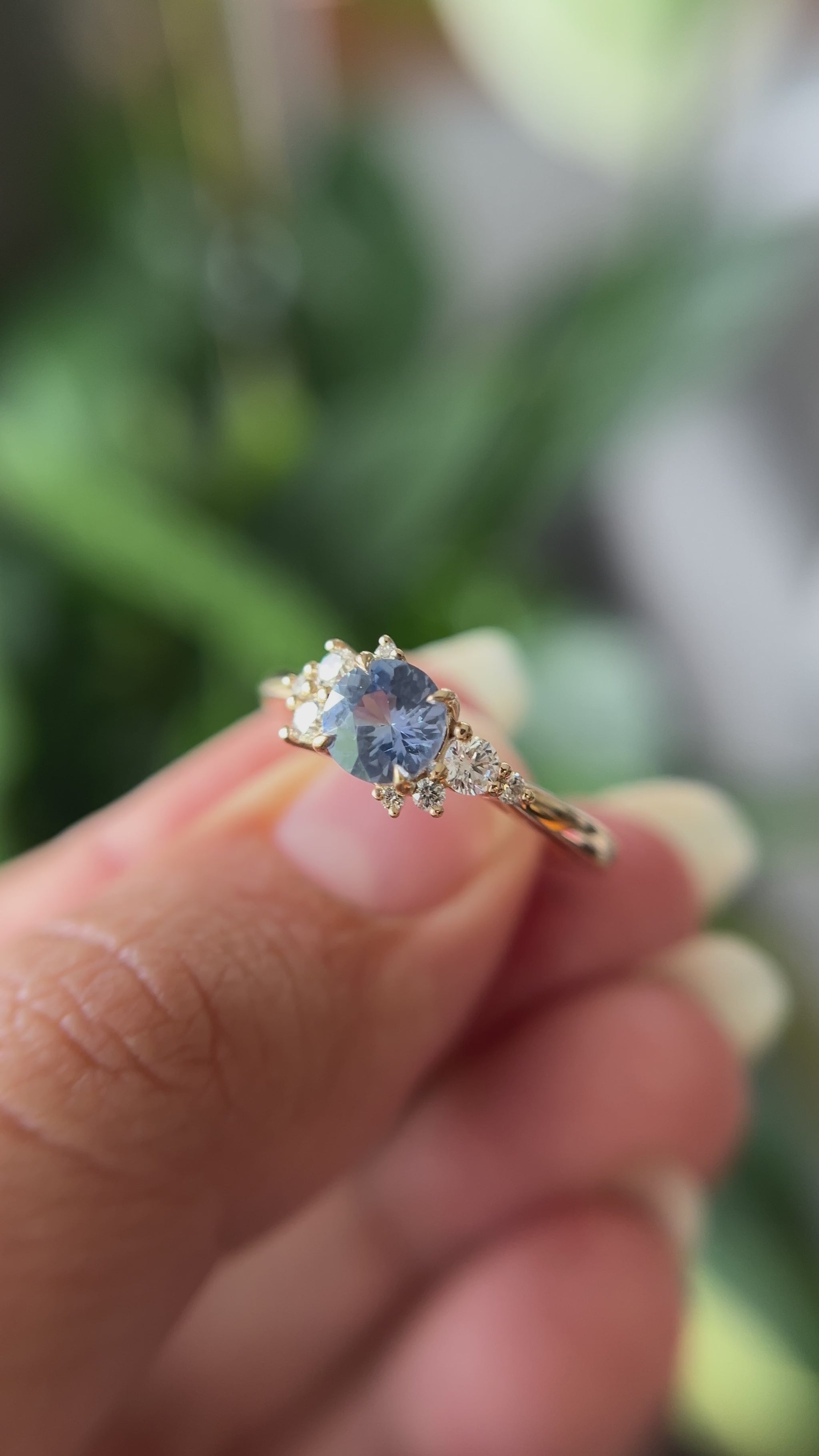 0.82ct round unheated cornflower blue Sri Lankan sapphire and diamond cluster engagement ring in 14k yellow gold, held in hands showing stone colour and asymmetric diamond setting in motion