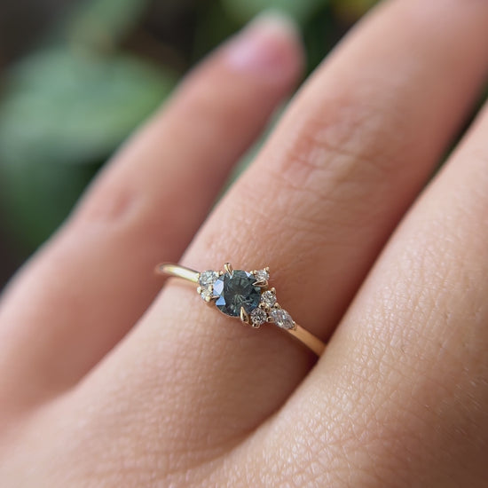 0.38ct round teal sapphire and natural diamond constellation engagement ring in 14k yellow gold, worn on finger showing gemstone colour and diamond cluster detail
