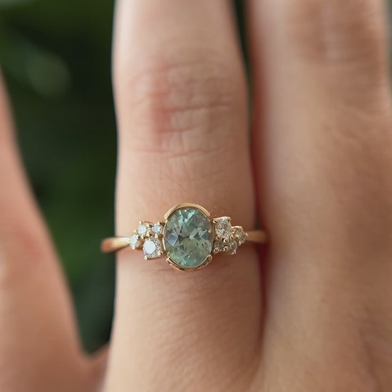 14k yellow gold ring, set with a teal-green oval sapphire from montana, and natural diamond accents. The ring is worn on a finger to show different angles of the ring, in front of a green blurry background.