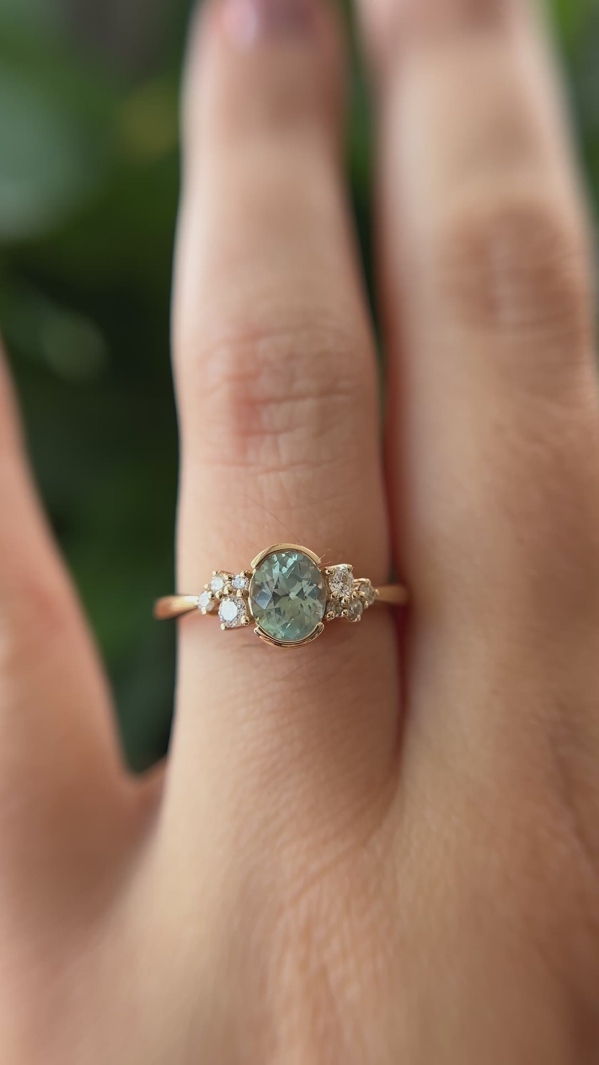 14k yellow gold ring, set with a teal-green oval sapphire from montana, and natural diamond accents. The ring is worn on a finger to show different angles of the ring, in front of a green blurry background.