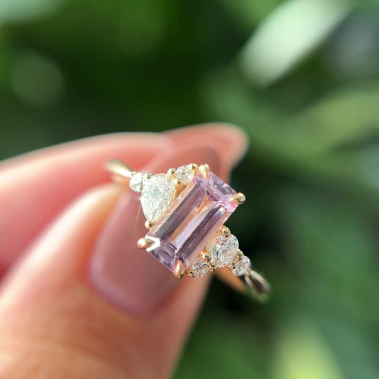 14k yellow gold ring, set with an emerald-cut light pink sapphire, and natural diamond accents. The ring is held between two fingers to show different angles of the ring, in front of a green blurry background.