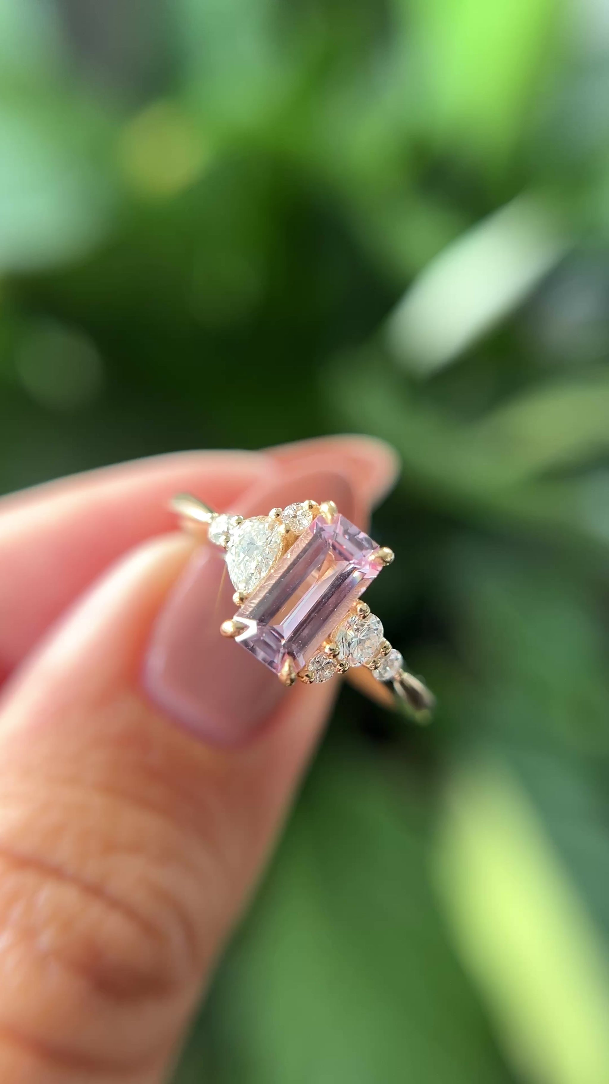 14k yellow gold ring, set with an emerald-cut light pink sapphire, and natural diamond accents. The ring is held between two fingers to show different angles of the ring, in front of a green blurry background.