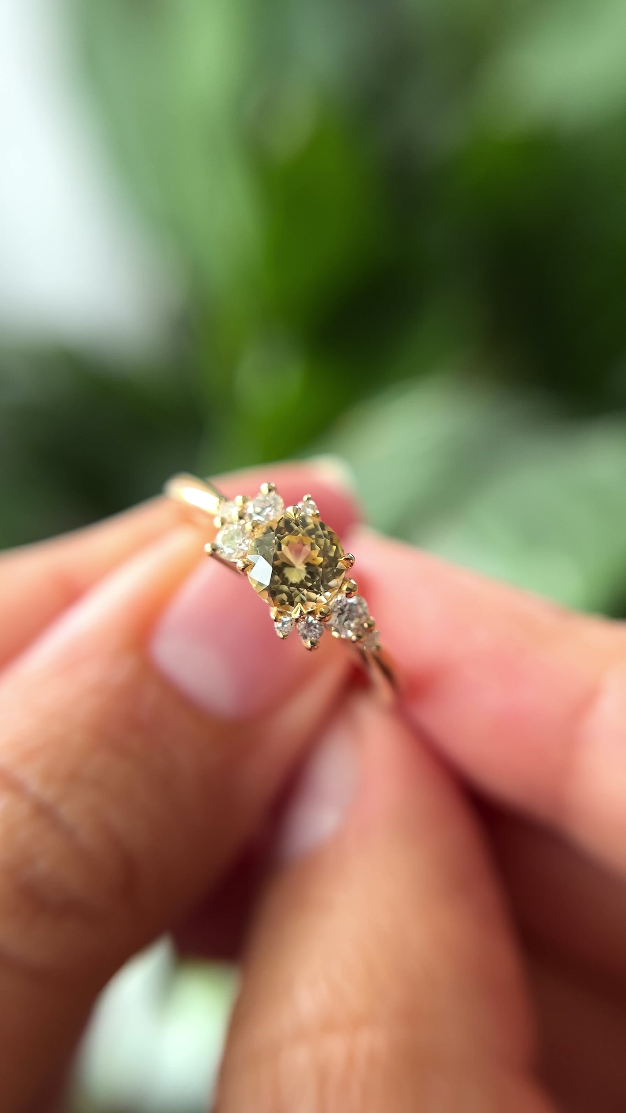 0.83ct round light yellow unheated Sri Lankan sapphire and diamond cluster engagement ring in 14k yellow gold, held in hands showing stone colour and asymmetric diamond setting in motion