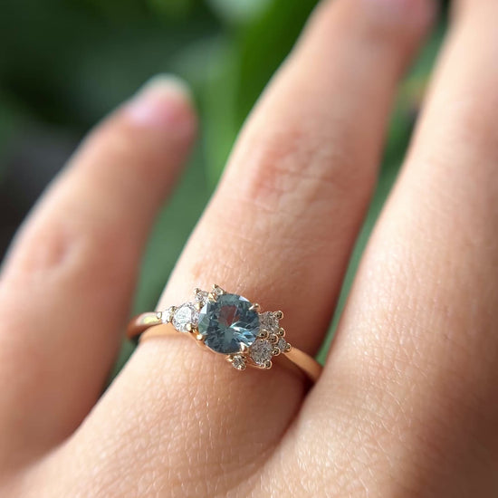 0.68ct round teal unheated sapphire and diamond cluster engagement ring in 14k yellow gold, worn on hand showing stone colour and asymmetric diamond setting in motion