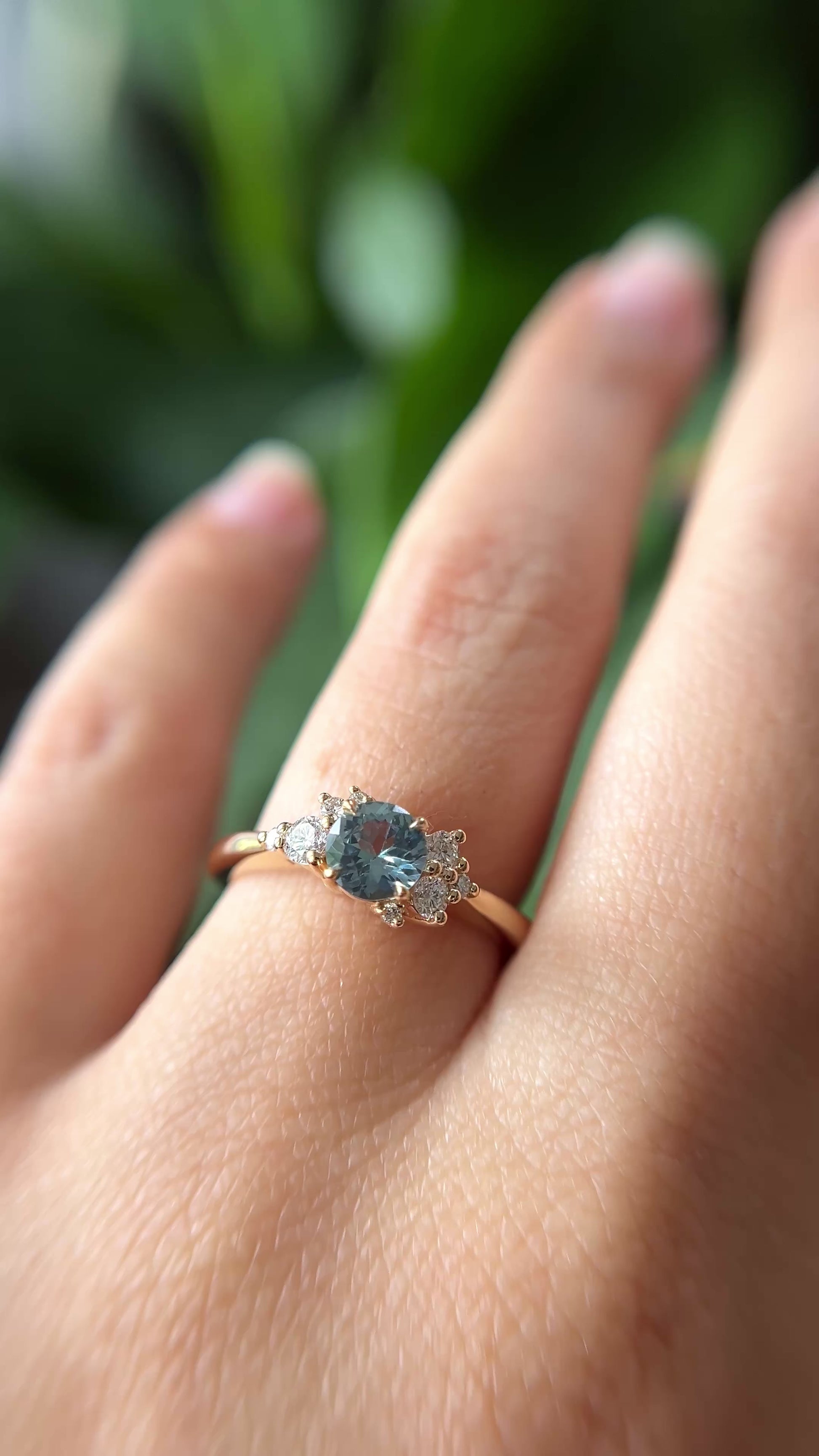 0.68ct round teal unheated sapphire and diamond cluster engagement ring in 14k yellow gold, worn on hand showing stone colour and asymmetric diamond setting in motion