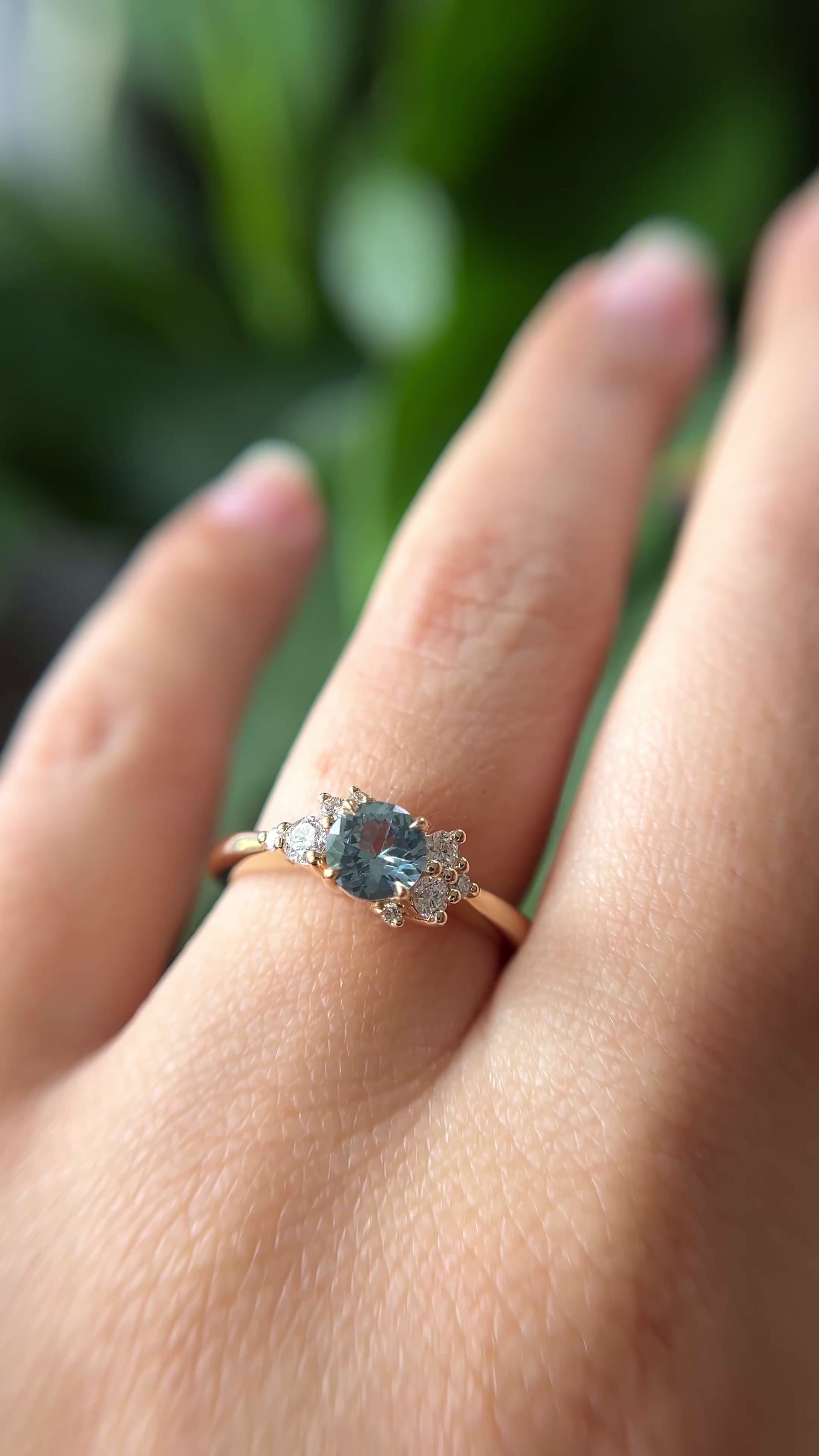0.68ct round teal unheated sapphire and diamond cluster engagement ring in 14k yellow gold, worn on hand showing stone colour and asymmetric diamond setting in motion