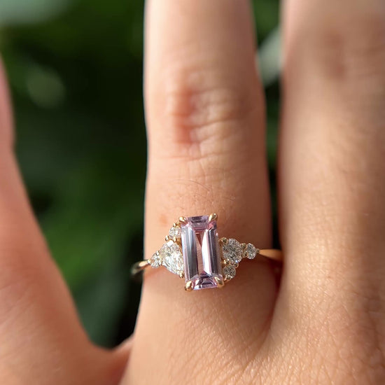 14k yellow gold ring, set with an emerald-cut light pink sapphire and natural diamond accents. The ring is worn on a finger to show different angles of the ring, in front of a green blurry background.