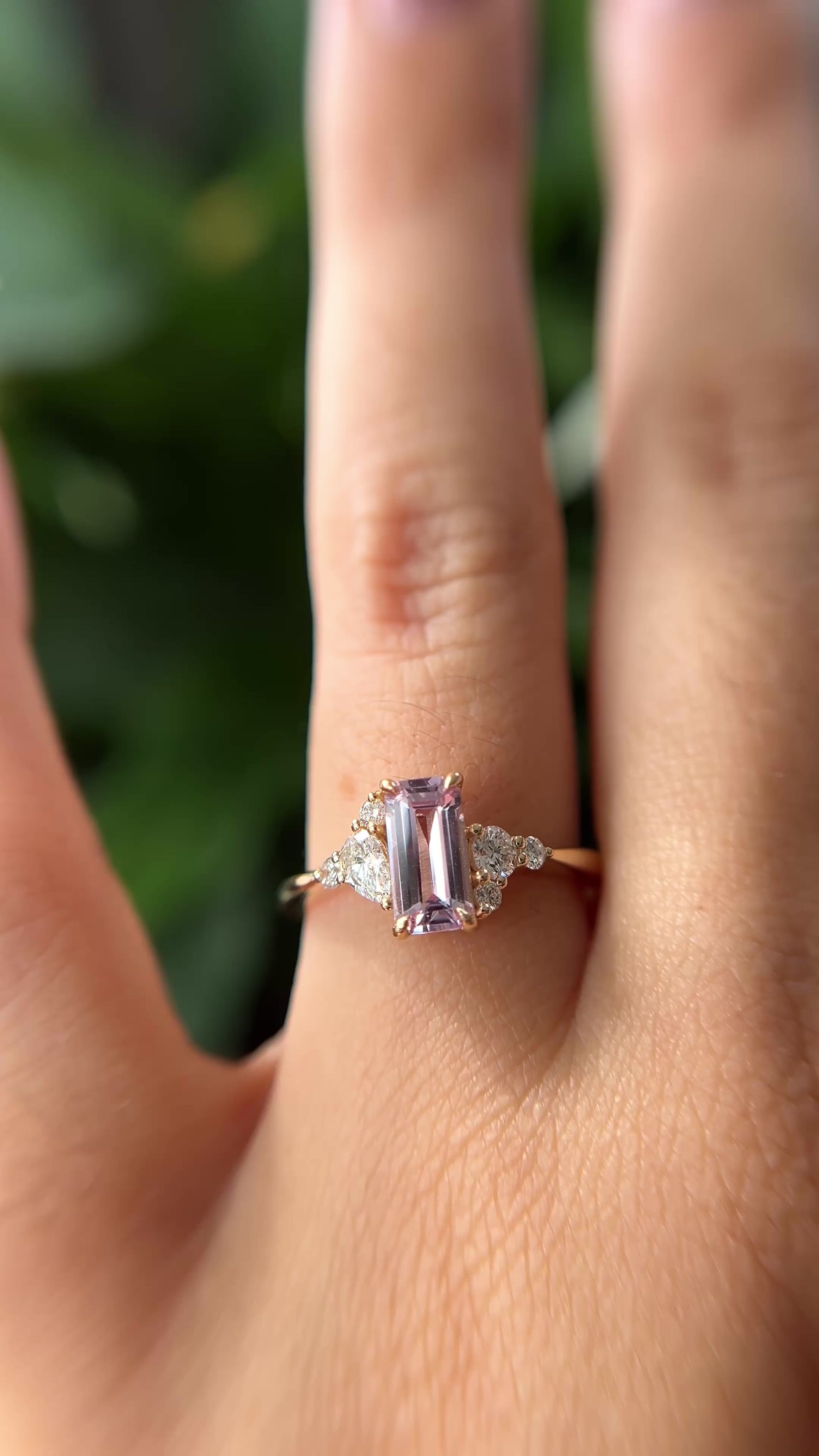 14k yellow gold ring, set with an emerald-cut light pink sapphire and natural diamond accents. The ring is worn on a finger to show different angles of the ring, in front of a green blurry background.