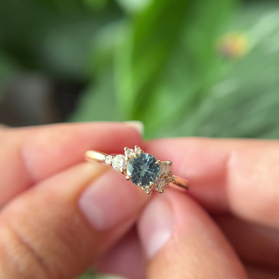 0.68ct round teal unheated sapphire and diamond cluster engagement ring in 14k yellow gold, held in hands showing stone colour and asymmetric diamond setting in motion