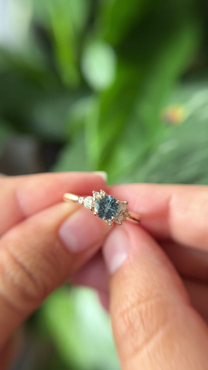 0.68ct round teal unheated sapphire and diamond cluster engagement ring in 14k yellow gold, held in hands showing stone colour and asymmetric diamond setting in motion