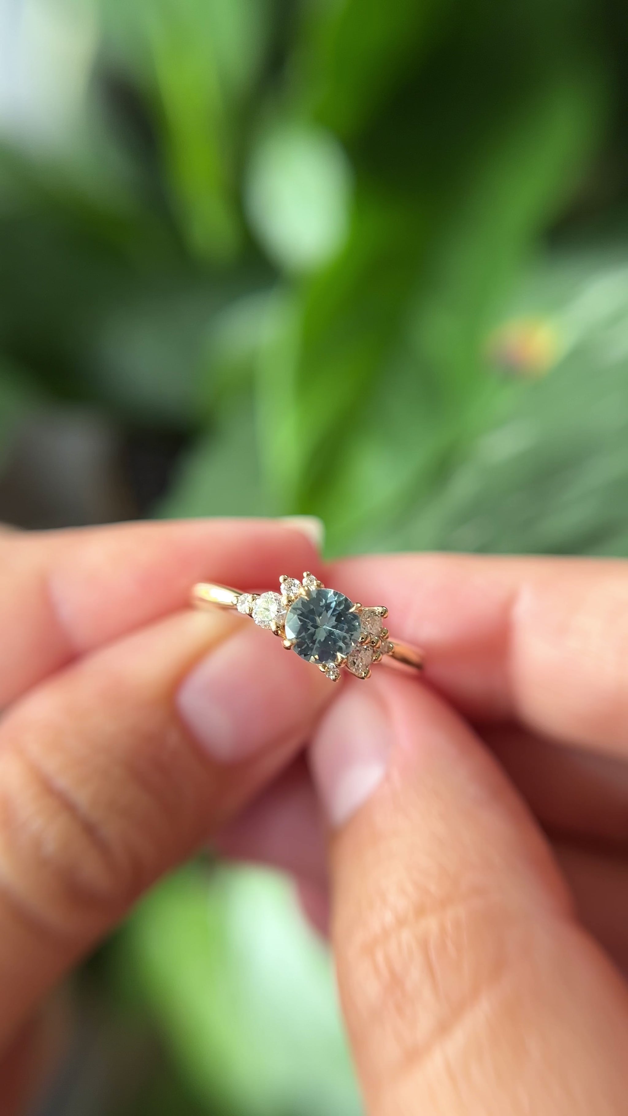 0.68ct round teal unheated sapphire and diamond cluster engagement ring in 14k yellow gold, held in hands showing stone colour and asymmetric diamond setting in motion