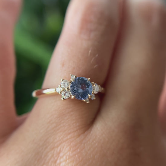 0.82ct round unheated cornflower blue Sri Lankan sapphire and diamond cluster engagement ring in 14k yellow gold, worn on hand showing stone colour and asymmetric diamond setting in motion
