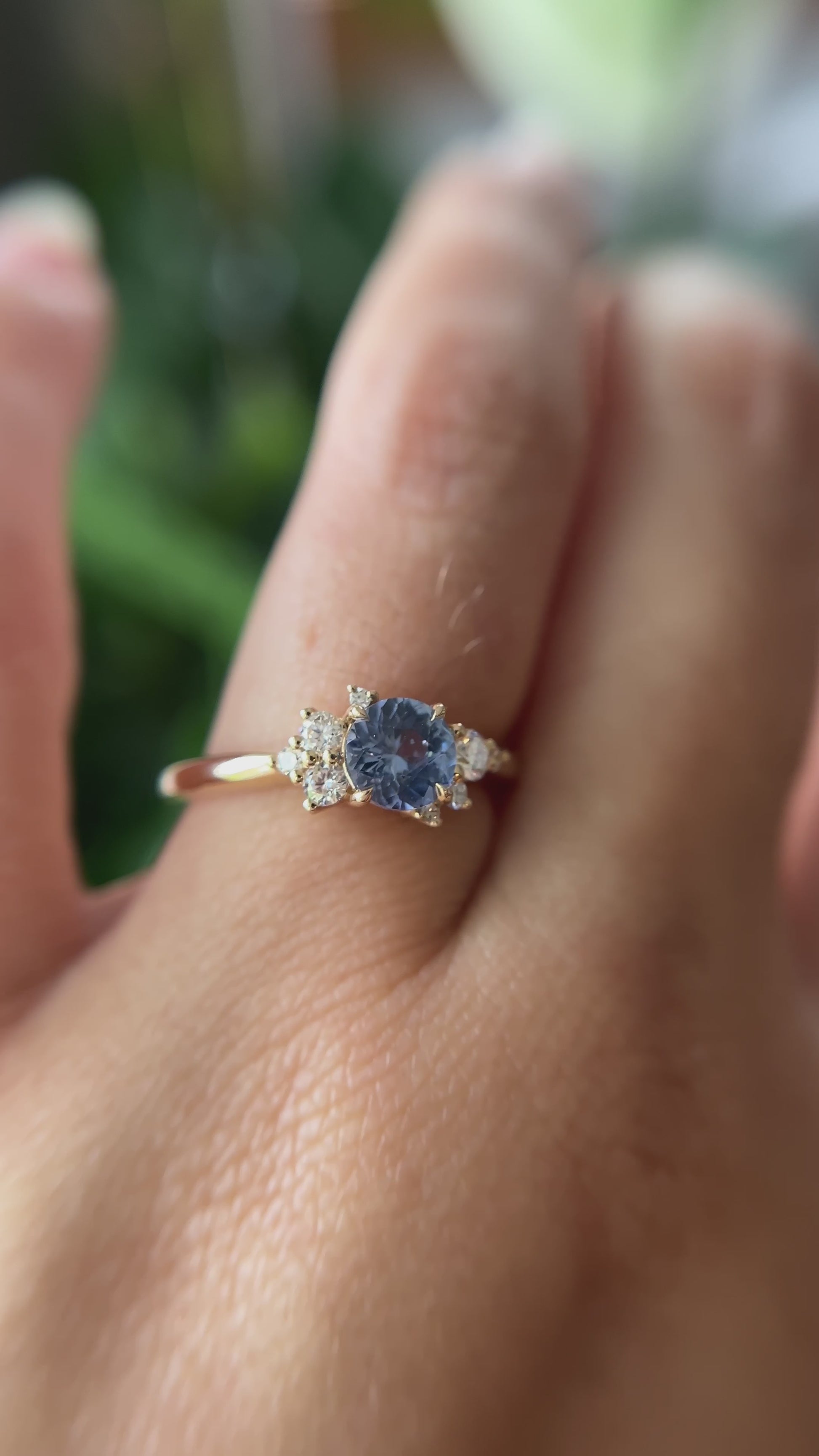 0.82ct round unheated cornflower blue Sri Lankan sapphire and diamond cluster engagement ring in 14k yellow gold, worn on hand showing stone colour and asymmetric diamond setting in motion