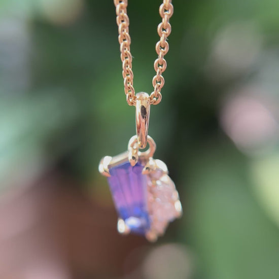 14k yellow gold pendant, set with a bi-color pink to blue sapphire and natural diamond accents. The pendant is hanging from a chain, showing the colors in different angles, in front of a green blurry background