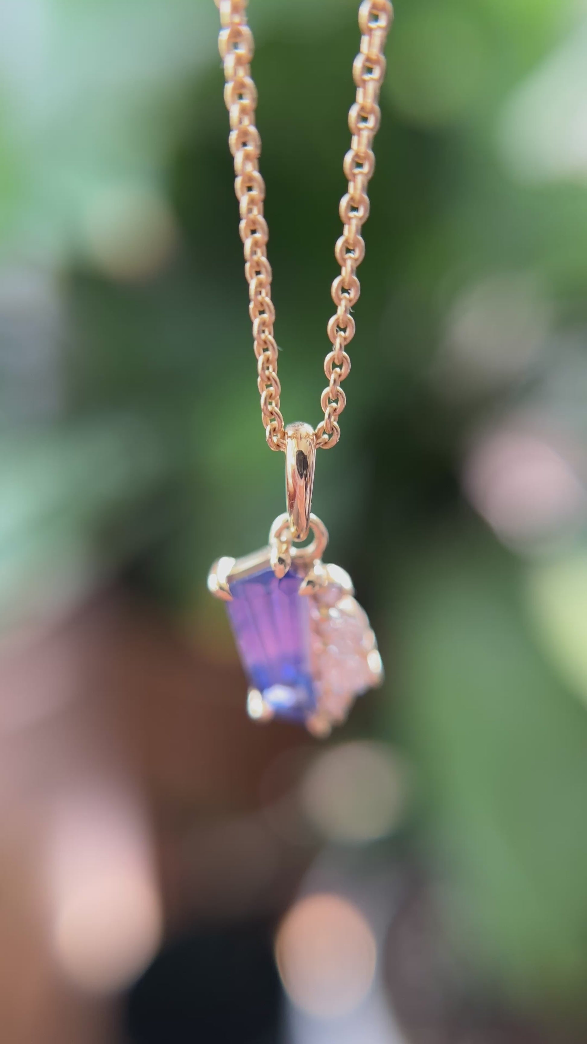 14k yellow gold pendant, set with a bi-color pink to blue sapphire and natural diamond accents. The pendant is hanging from a chain, showing the colors in different angles, in front of a green blurry background