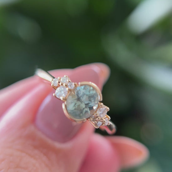 14k yellow gold ring, set with a teal-green oval sapphire from montana, and natural diamond accents. The ring is held between two fingers to show different angles of the ring, in front of a green blurry background.