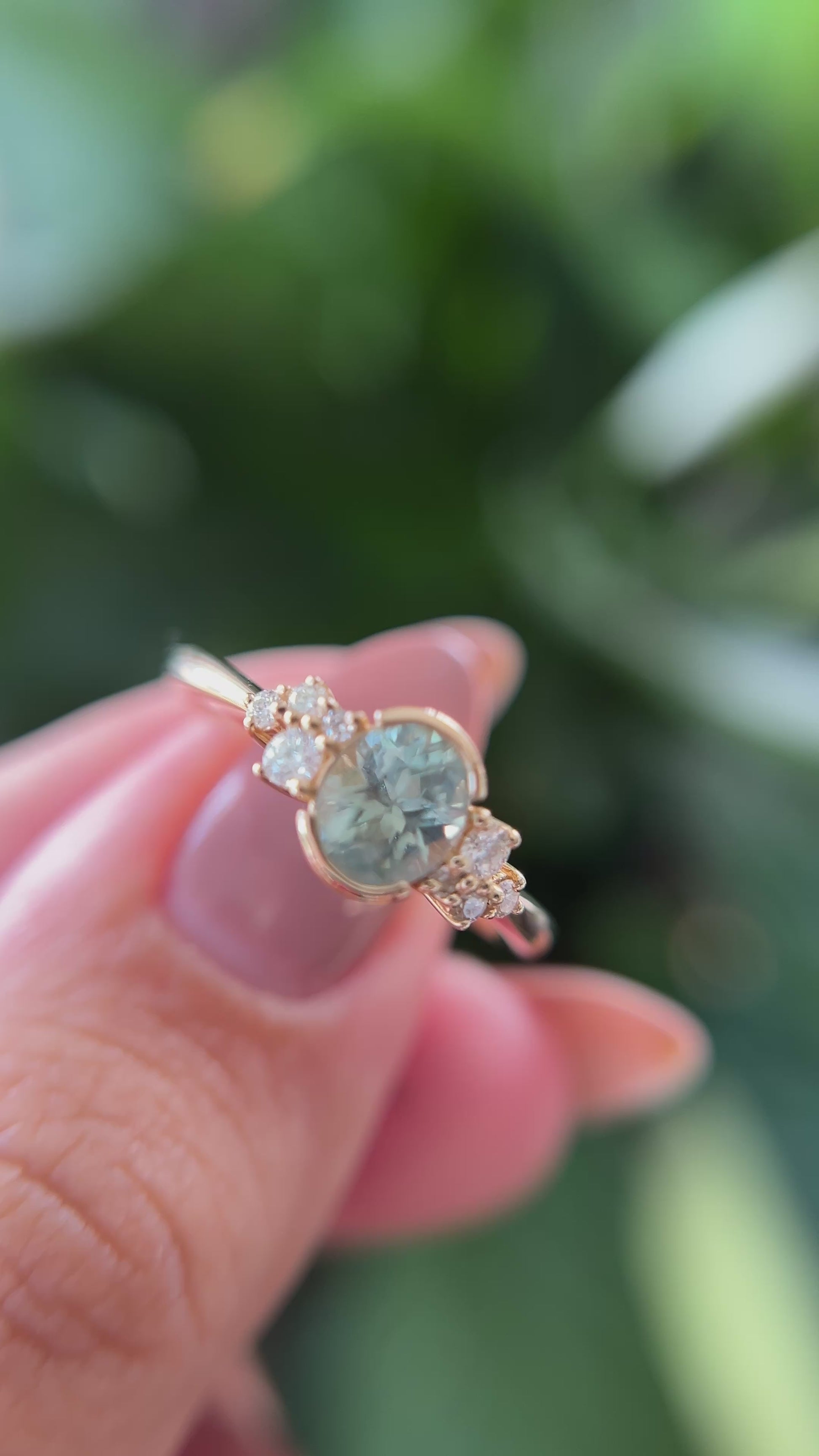 14k yellow gold ring, set with a teal-green oval sapphire from montana, and natural diamond accents. The ring is held between two fingers to show different angles of the ring, in front of a green blurry background.