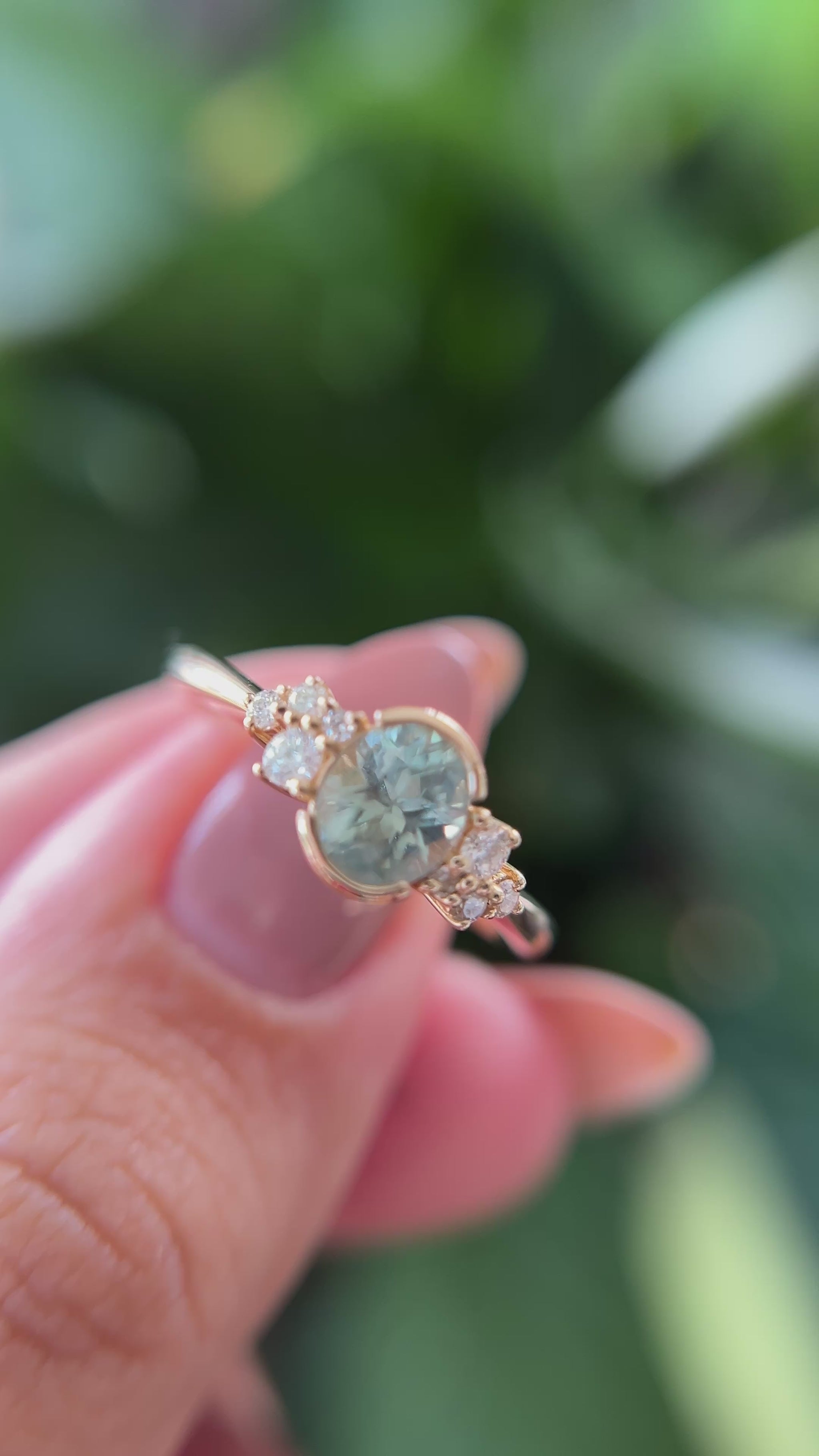 14k yellow gold ring, set with a teal-green oval sapphire from montana, and natural diamond accents. The ring is held between two fingers to show different angles of the ring, in front of a green blurry background.