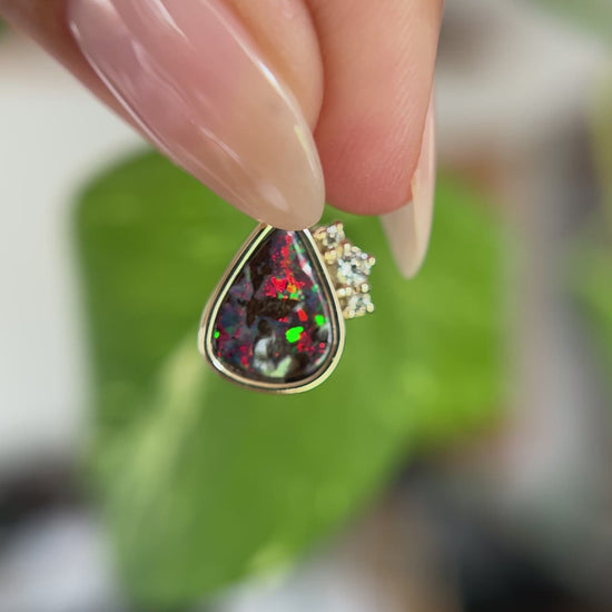 14k yellow gold pendant, bezel set with pear-shaped boulder opal from Australia and claw set diamond accents. The pendant is held between two fingers to show different angles and play of color. In front of a green blurry background.