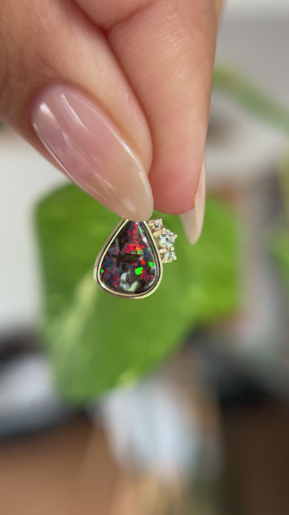 14k yellow gold pendant, bezel set with pear-shaped boulder opal from Australia and claw set diamond accents. The pendant is held between two fingers to show different angles and play of color. In front of a green blurry background.