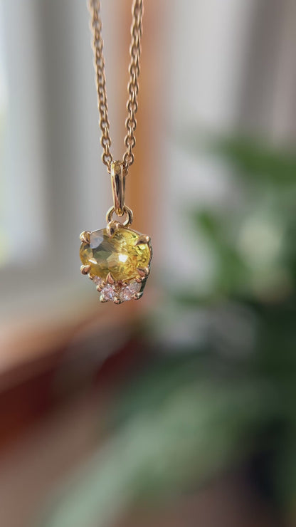 14k yellow gold pendant, set with a oval golden beryl and natural diamond accents. The pendant is hanging from a chain, in front of a green blurry background