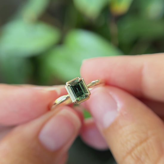 1.05ct emerald-cut forest green Madagascar sapphire solitaire engagement ring in 14k yellow gold, held in hands showing gemstone colour and faceting
