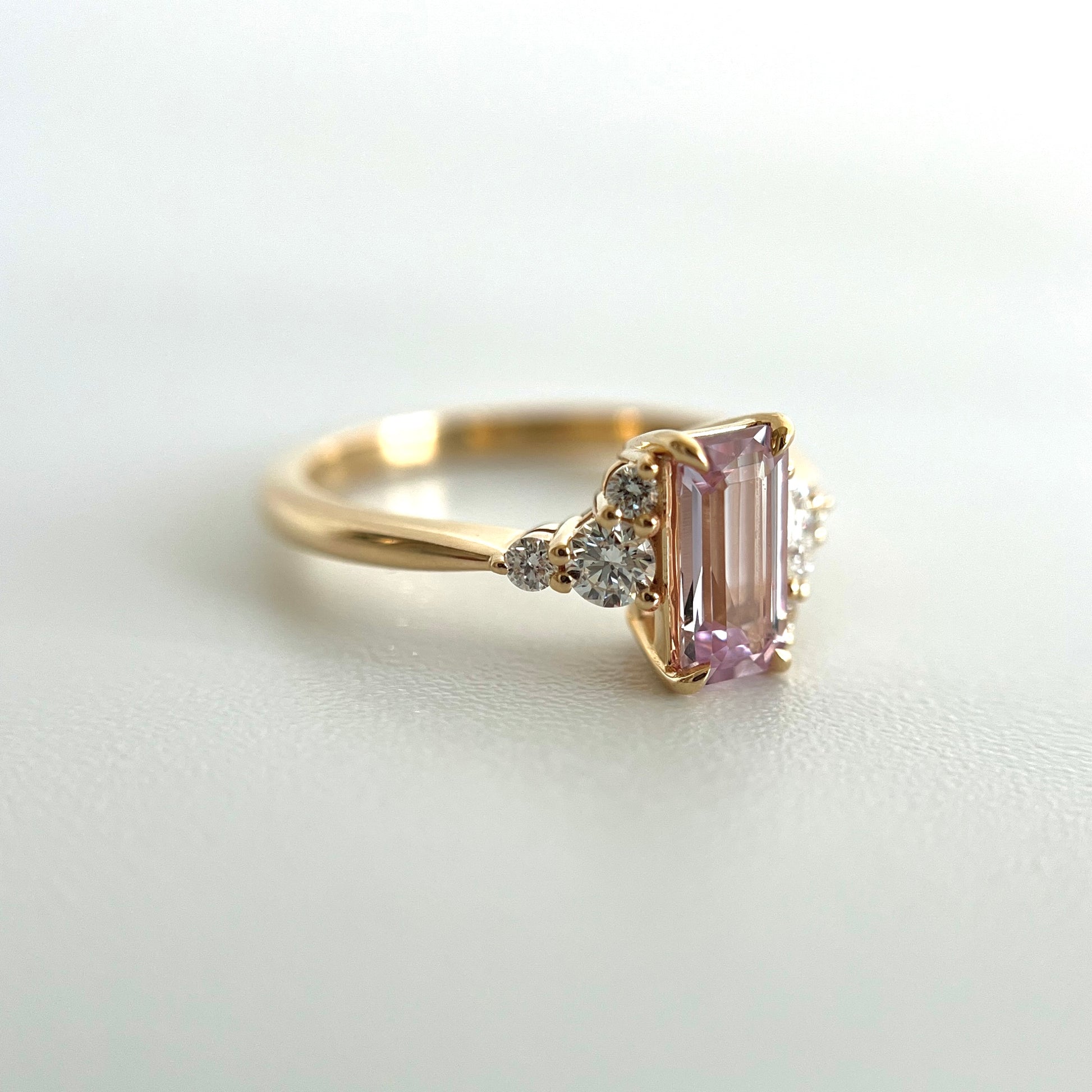 Side view of a 14k yellow gold ring, with an eagle-claw set emerald-cut light pink sapphire and diamond accents. The photo focuses on the setting details on the 3 round side diamonds on the left. 