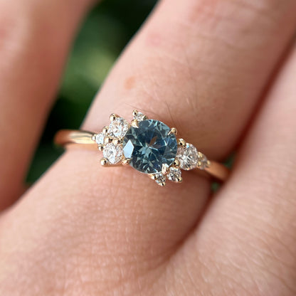 Gros plan de la bague de fiançailles en saphir sarcelle et diamants en or jaune 14k, portée au doigt montrant la couleur sarcelle de la pierre et le détail du serti