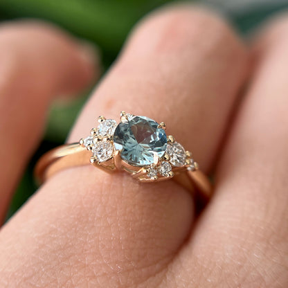 Bague de fiançailles en saphir sarcelle et diamants en or jaune 14k, portée au doigt montrant la couleur de la pierre et le détail asymétrique des diamants