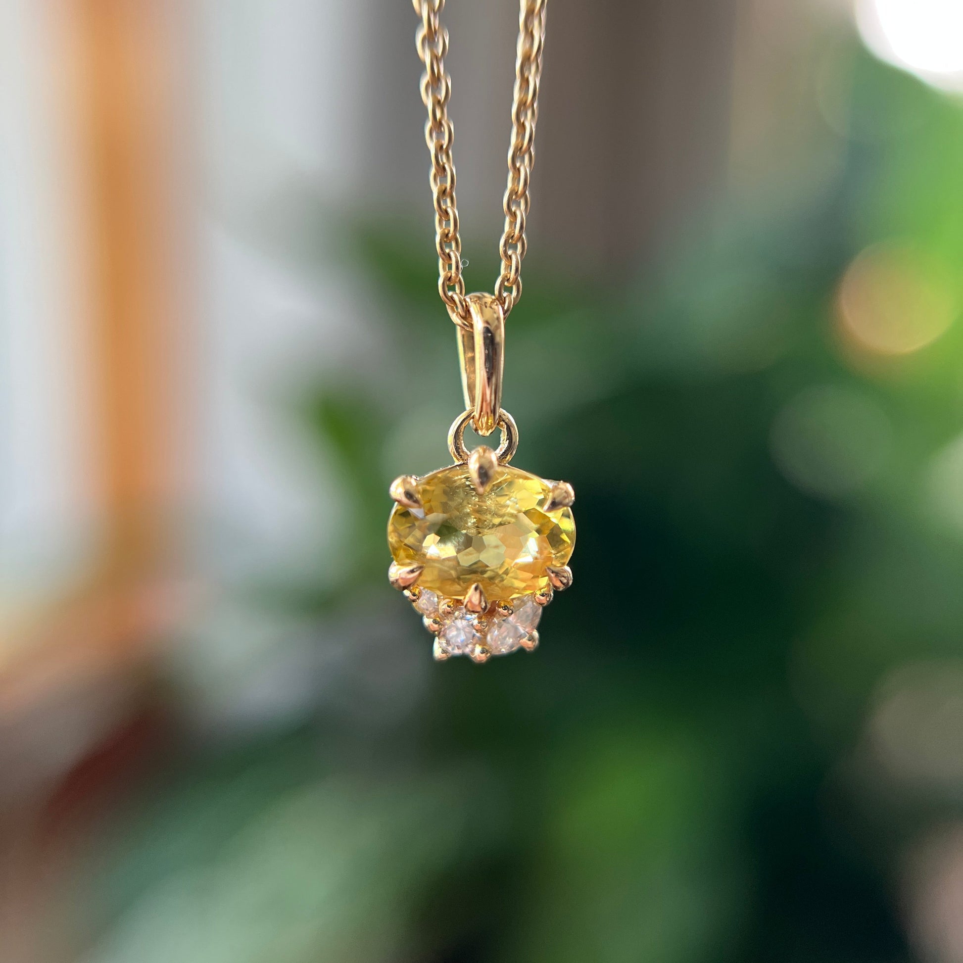 A gold pendant necklace featuring a oval yellow heliodore with small natural diamonds accent stones, hanging against a softly blurred green and brown background.